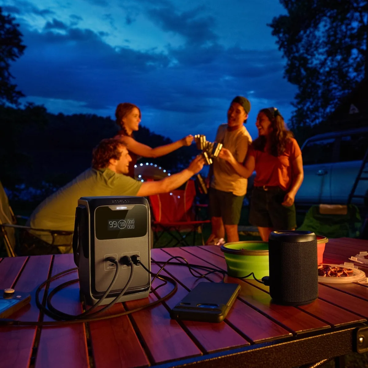 An EcoFlow TRAIL 200 DC on a table at a campsite charging a phone and a speaker while friends relax