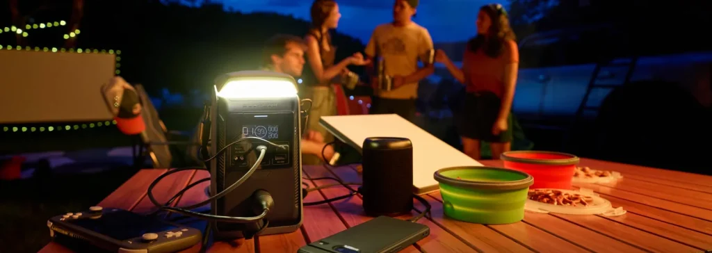 EcoFlow TRAIL Plus 300 DC з увімкненим ліхтарем на столі в кемпінгу, поруч колонка і телефони